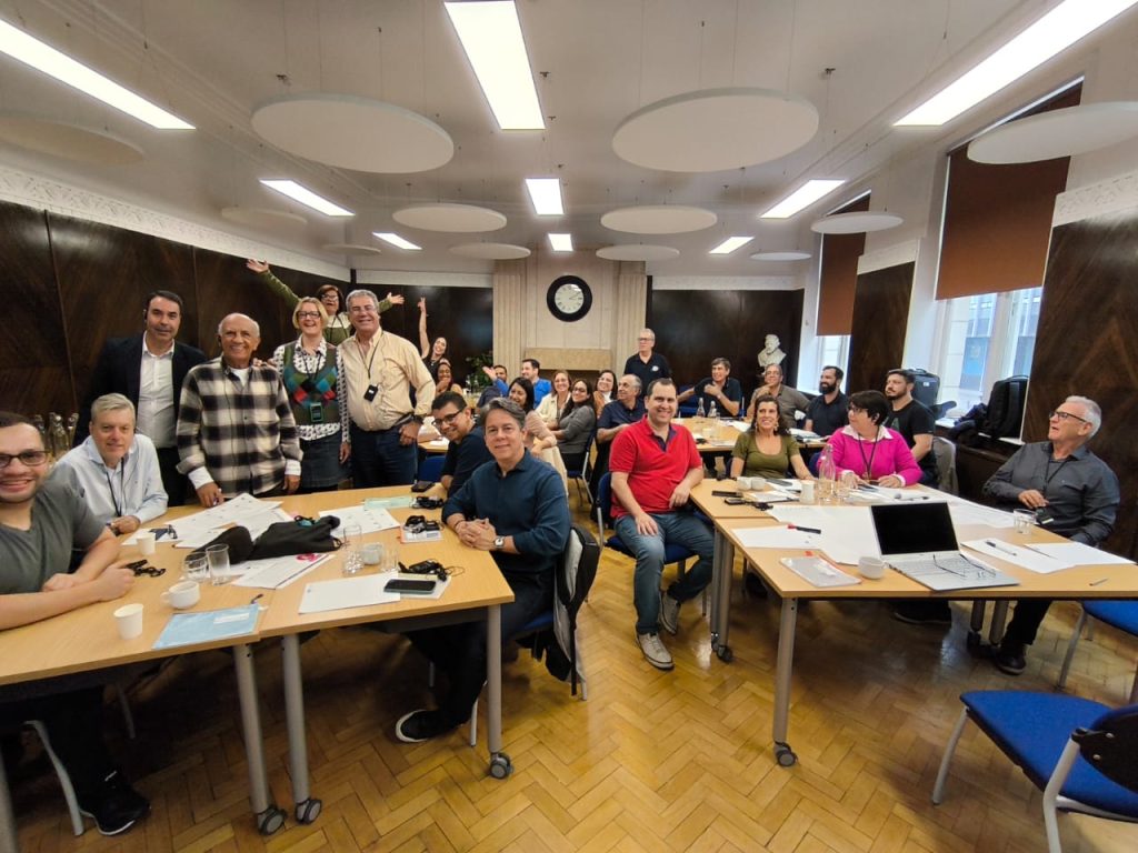 Uma sala de reuniões cheia de pessoas, distribuídas em várias mesas. Há papéis, laptops e bebidas sobre as mesas. A sala tem piso de madeira, iluminação moderna e grandes janelas que permitem a entrada de luz natural. O grupo é composto por homens e mulheres, todos aparentando estar felizes e engajados, possivelmente participando de um workshop ou treinamento. Algumas pessoas estão em pé, outras sentadas, e no fundo da sala há um relógio grande e uma estátua branca. O ambiente é descontraído e positivo.