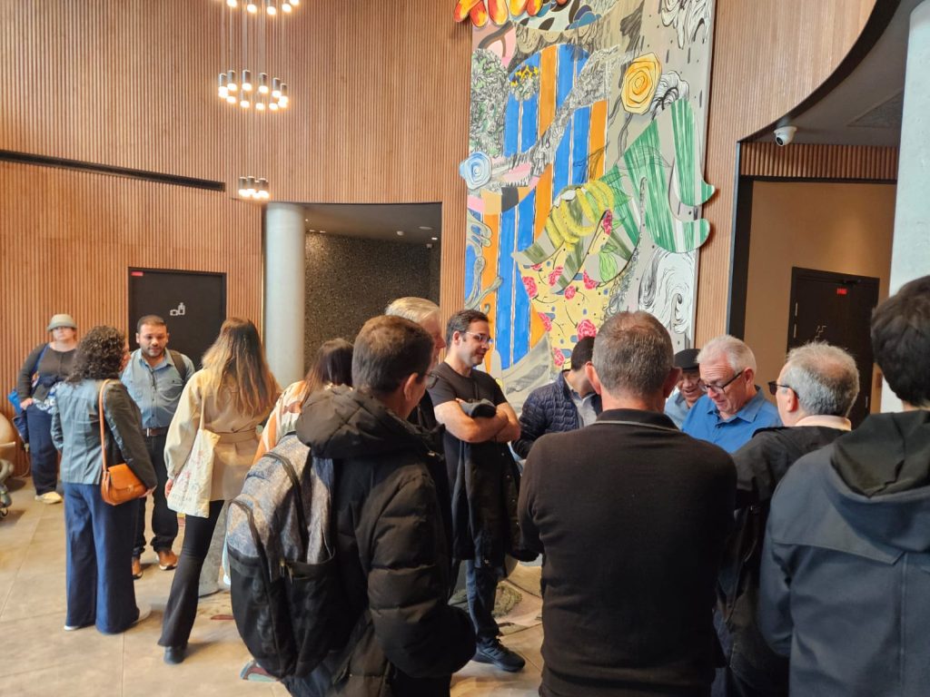 Grupo diversificado de pessoas interagindo em um espaço interno decorado com painéis de madeira e um mural colorido, discutindo ativamente em um ambiente informal