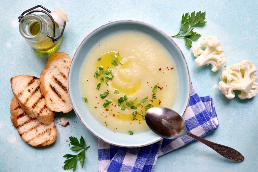 Tigela de sopa cremosa de couve-flor com batata, decorada com azeite e salsinha picada, ao lado de fatias de pão grelhado e floretes de couve-flor