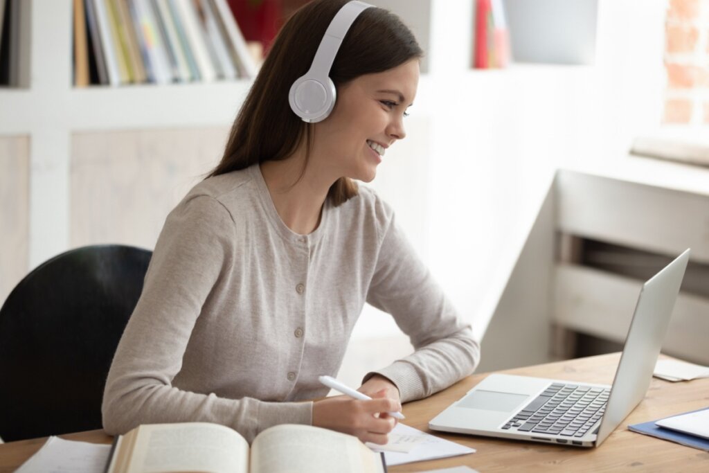 modelo feminino simula, sorridente, estar se preparando para o Enem ouvindo matéria diante de um laptop sobre mesa com livro e caderno de anotação 