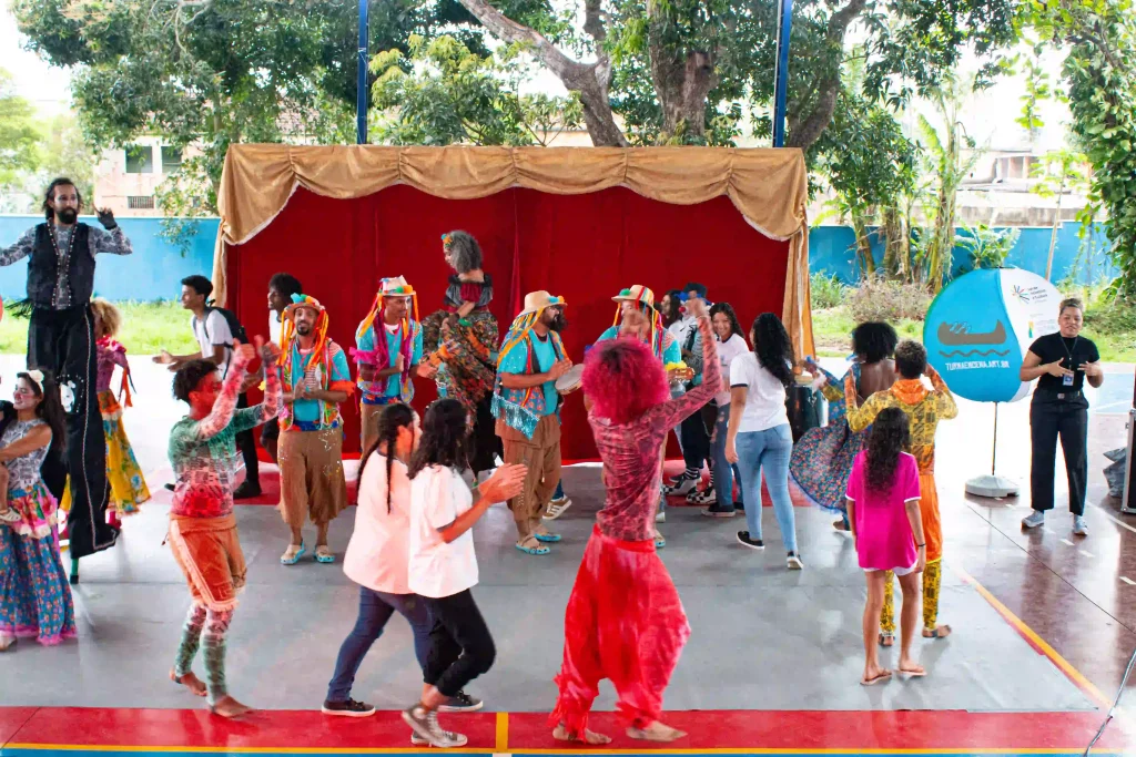 A imagem retrata uma interação festiva entre artistas circenses e o público. Em frente a um pano de fundo vermelho, vários artistas vestidos com roupas coloridas e vibrantes, incluindo um homem em pernas de pau, dançam e se divertem ao lado de participantes da comunidade. O cenário sugere um ambiente descontraído e de inclusão, onde o público, composto por pessoas de diversas idades, participa ativamente das atividades. Ao fundo, há árvores e uma placa que indica um projeto comunitário ou cultural, reforçando o caráter de celebração popular e integração social.