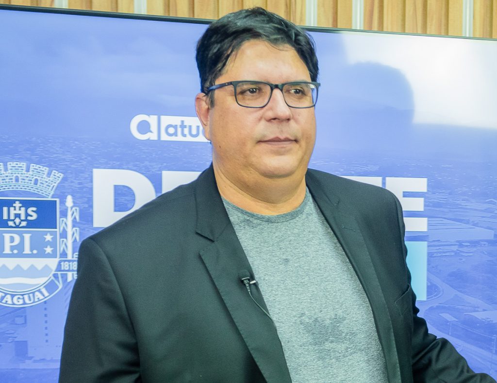 o jornalista Marcelo Godinho é fotografado usando  costume preto e blusa cinza a frente de uma TV que mostra o brasão heráldico da cidade de Itaguaí ao lado de uma palavra encoberta pelo fotografado  