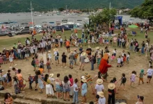Roda formada por dezenas de pessoas — crianças, jovens e adultos — participando de atividades culturais ao ar livre em Paraty, com o mar e barcos ao fundo, durante o 1º Festival de Cultura Caiçara da Costa Verde.
