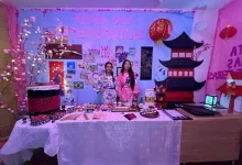 Duas estudantes vestidas com quimonos posam em frente a um estande temático da cultura japonesa, com decoração de cerejeiras, lanternas, maquete de pagode e mesa com objetos e comidas típicas, durante a Feira Mundo do Colégio Educandário São José REALIZADA EM 2024