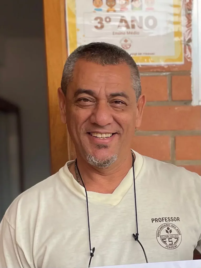 Professor do Colégio Educandário São José sorri para a foto; ele veste camiseta da escola com o logotipo da instituição e está em frente a um mural com a indicação de ‘3º ano do Ensino Médio’