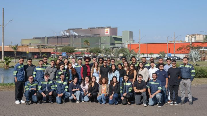 Grupo de trabalhadores e colaboradores posa para foto em frente a complexo industrial com galpões e chaminés ao fundo, em dia ensolarado.